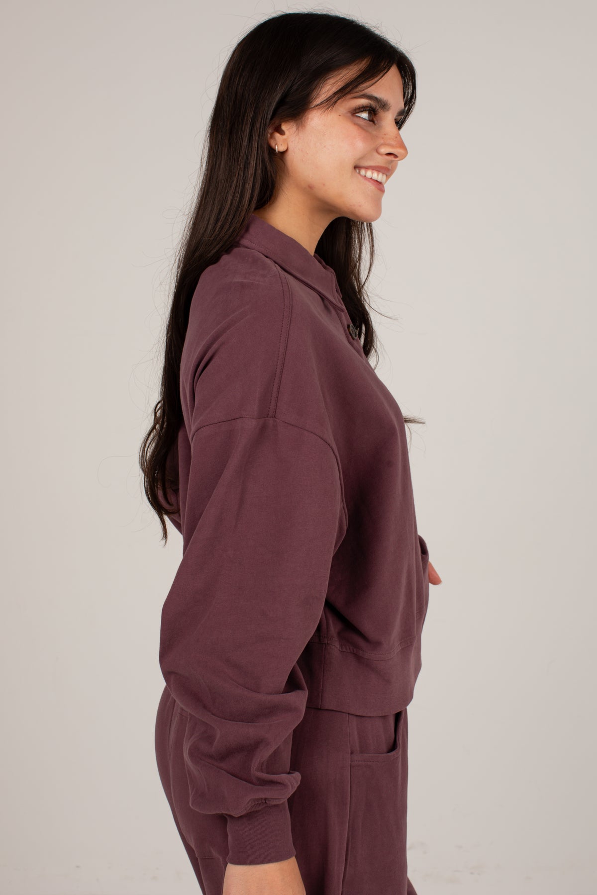 A woman with long dark hair is shown in profile wearing a matching dusty plum-brown lounge set consisting of a long-sleeve collared top and pants, against a plain light gray studio background.