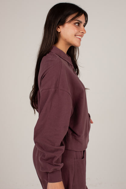 A woman with long dark hair is shown in profile wearing a matching dusty plum-brown lounge set consisting of a long-sleeve collared top and pants, against a plain light gray studio background.