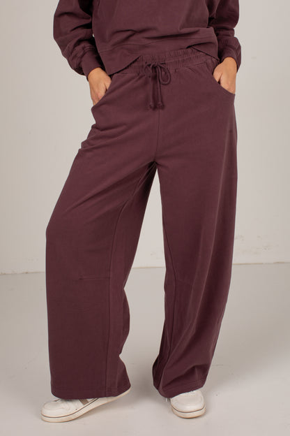 A person wearing loose, wide-leg burgundy pants with a drawstring waistband and side pockets, paired with white sneakers, shown from the waist down against a plain light gray studio background.