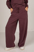 A person wearing loose, wide-leg burgundy pants with a drawstring waistband and side pockets, paired with white sneakers, shown from the waist down against a plain light gray studio background.