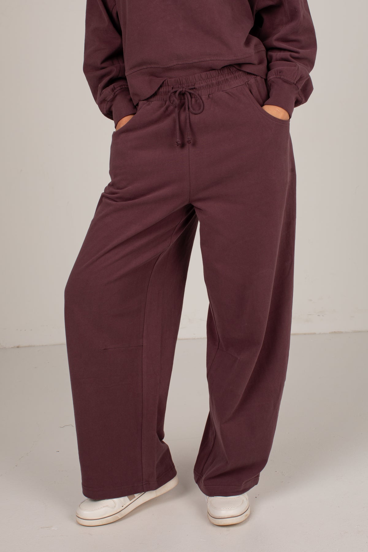 A person wearing loose, wide-leg drawstring sweatpants in a deep plum-brown color, paired with a matching sweatshirt and white sneakers, shown from the chest down against a plain studio background.