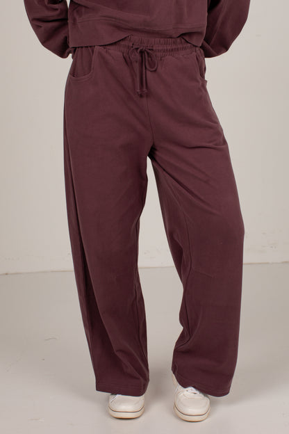 A person wearing loose, wide-leg drawstring sweatpants in a deep plum-burgundy color, paired with white sneakers, shown from the waist down against a plain off-white studio background.