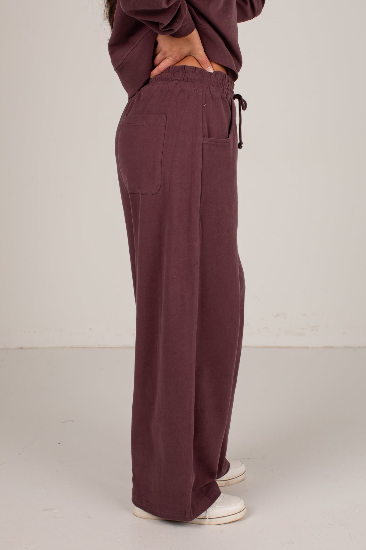 A person shown from the waist down wearing loose, wide-leg drawstring pants in a muted plum-brown color, paired with white sneakers against a plain off-white studio background.