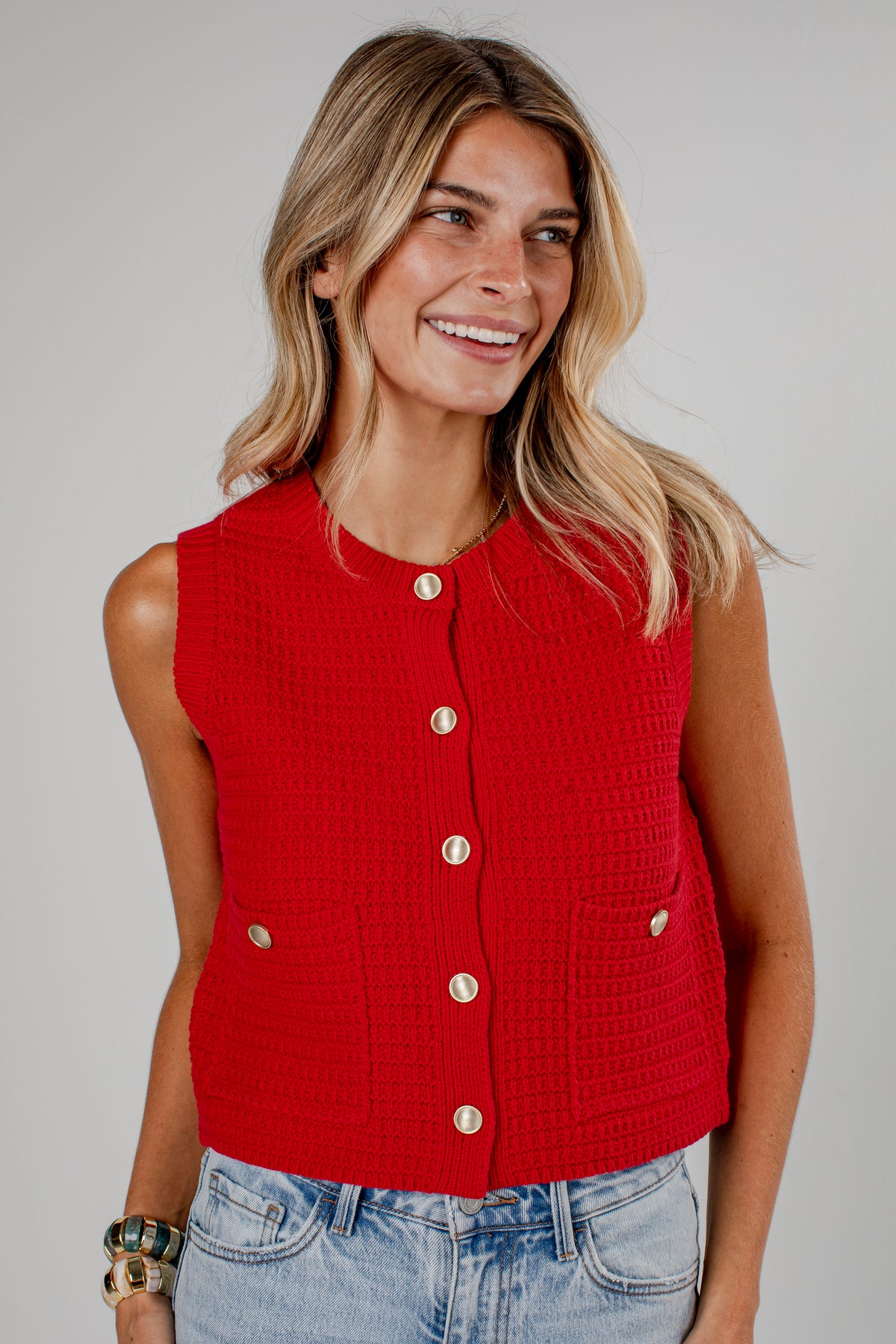 A woman wearing a bright red sleeveless cropped knit vest with gold-tone buttons and front pockets, paired with light-wash blue jeans, against a solid light gray studio background.