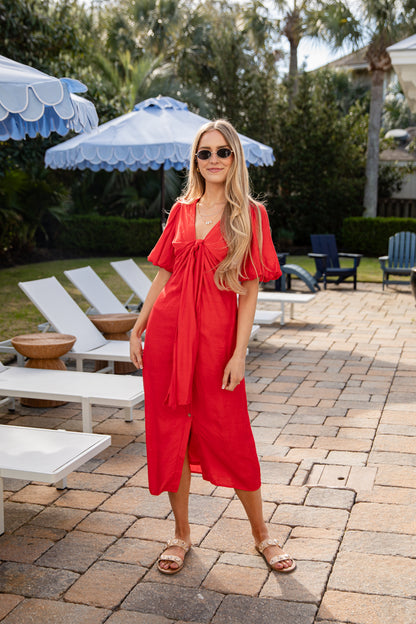 Woman in a red dress standing on a patio with lounge chairs and umbrellas in the background.