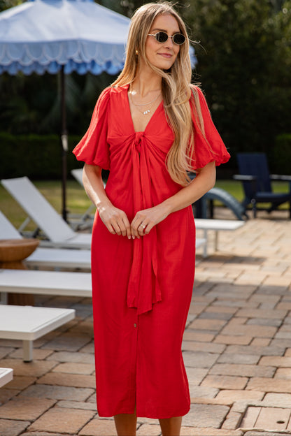 Woman in a red dress standing outdoors with lounge chairs and an umbrella in the background