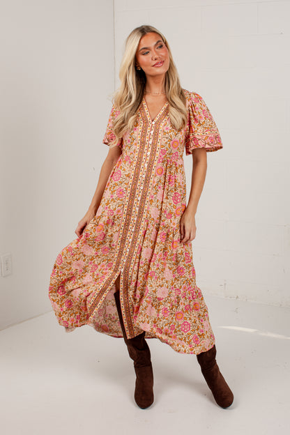 A woman modeling a short-sleeve bohemian-style maxi dress with a pink and orange floral pattern and a gold patterned center panel, paired with brown boots, in a white studio setting.