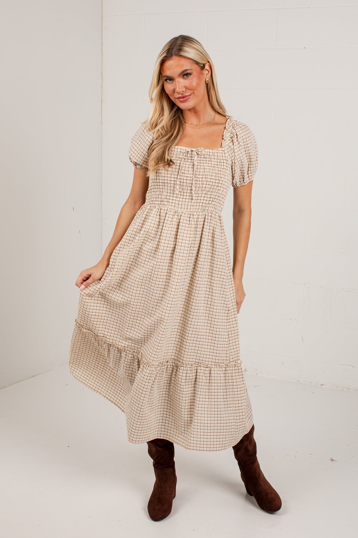 A woman modeling a beige and white gingham tiered midi dress with puff sleeves and a smocked bodice, paired with brown boots, in a white studio setting.