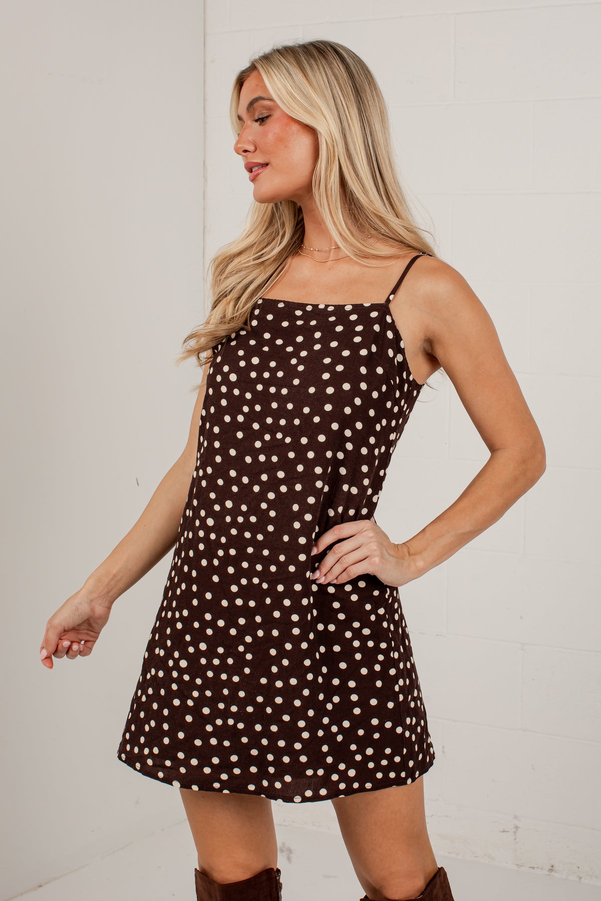 A blonde woman modeling a brown mini dress with a white polka dot pattern, featuring thin spaghetti straps and a square neckline, posed against a white brick wall.