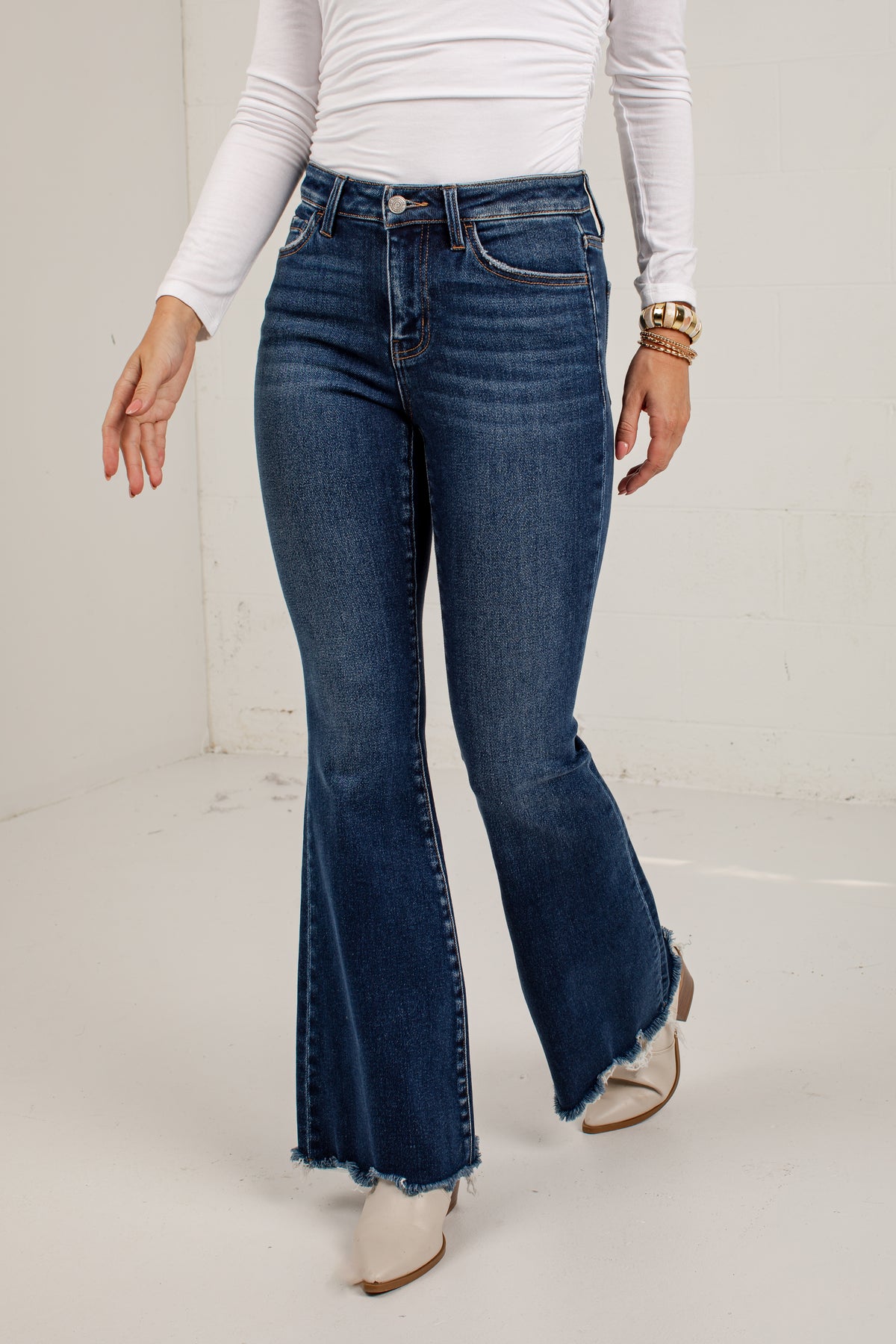 A model wearing dark blue high-waisted flare jeans with a frayed hem, paired with a white long-sleeve top and white pointed-toe heels, standing against a white studio wall.