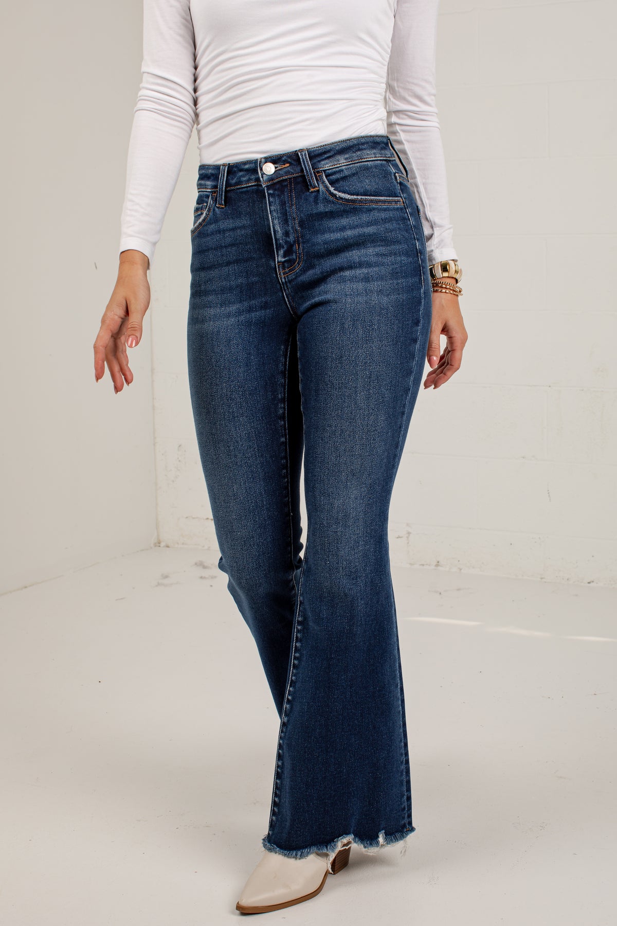 A model wearing dark blue flare jeans with a frayed hem, paired with a white long-sleeve top and white pointed-toe heeled boots, standing against a white studio wall.