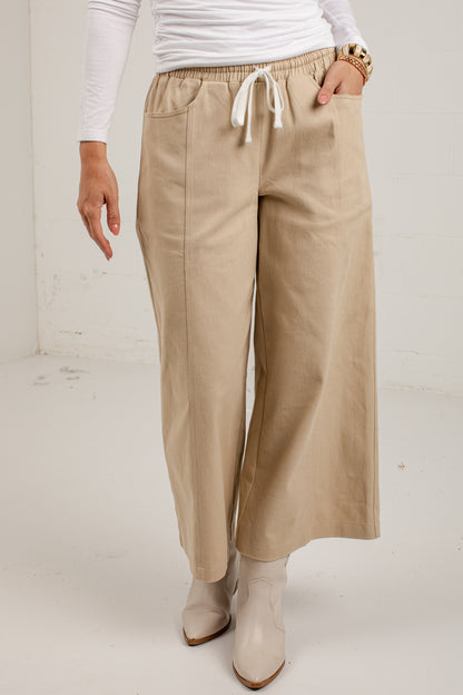 A person wearing beige wide-leg cropped pants with an elastic waistband and white drawstring, paired with a white long-sleeve top and beige ankle boots.