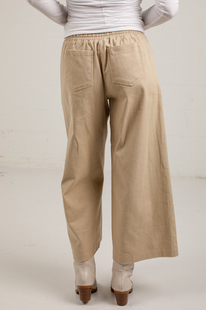 A person shown from the back wearing loose, wide-leg beige pants with an elastic waistband and back pockets, paired with a white ribbed long-sleeve top and light-colored heeled boots against a plain off-white studio background.