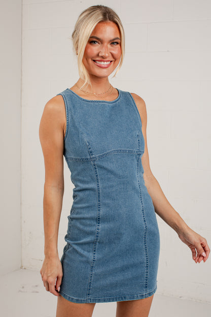 A woman wearing a sleeveless blue denim mini dress, standing against a white brick wall.