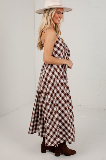 A woman wearing a brown and white gingham maxi dress with a tie-shoulder design, paired with a cream wide-brim hat and brown ankle boots, standing against a white studio wall.