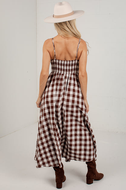 A woman shown from the back wearing a brown and cream gingham maxi dress with thin spaghetti straps, a smocked bodice, and a flared skirt. She is also wearing a cream wide-brim hat and brown suede ankle boots, standing against a white studio background.