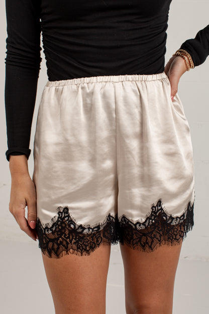 A person wearing satin shorts in a warm off-white/cream color with a black lace hem, paired with a black long-sleeve top. The photo is cropped from the chest down against a light gray background.