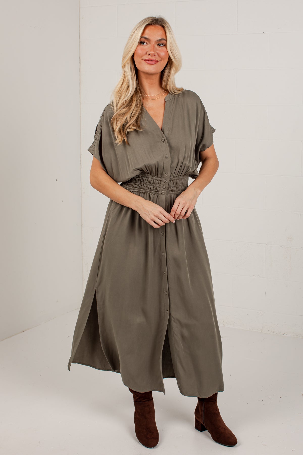 A woman modeling a short-sleeve, smocked-waist midi dress in a muted olive green color, paired with brown suede ankle boots, standing against a white studio wall.