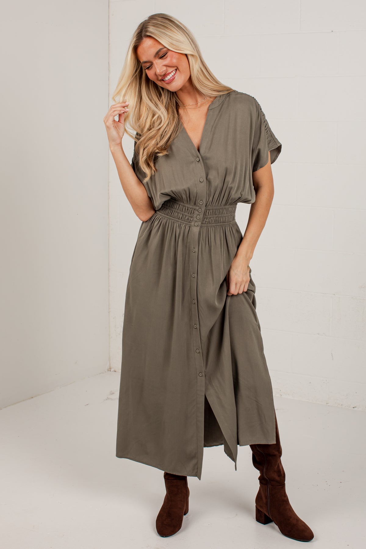A woman modeling a short-sleeve, button-front midi dress in a muted olive green color, paired with brown suede boots, standing against a white studio wall.