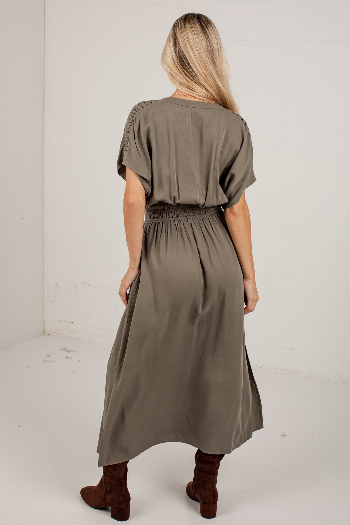 A woman viewed from behind wearing an olive green midi dress with short flutter sleeves, a gathered waist, and brown ankle boots, standing against a plain white wall.