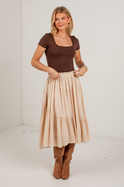 A woman modeling a tiered midi skirt in a light beige color, paired with a brown short-sleeve top and brown boots, standing against a plain white studio wall.