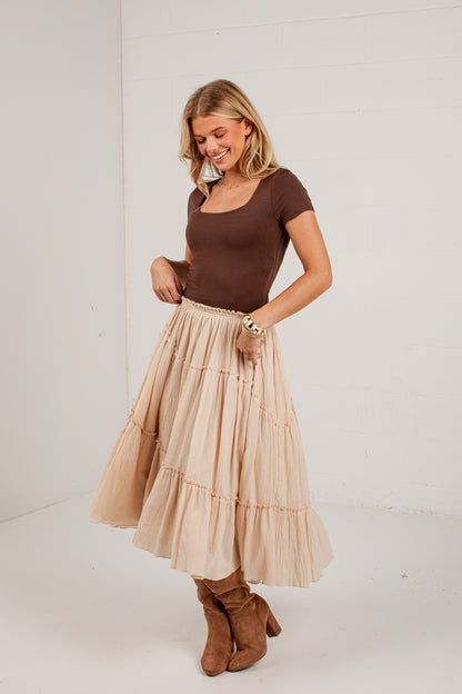 A woman modeling a brown short-sleeve scoop-neck top and a tiered beige maxi skirt, paired with brown suede boots, standing against a white brick wall.
