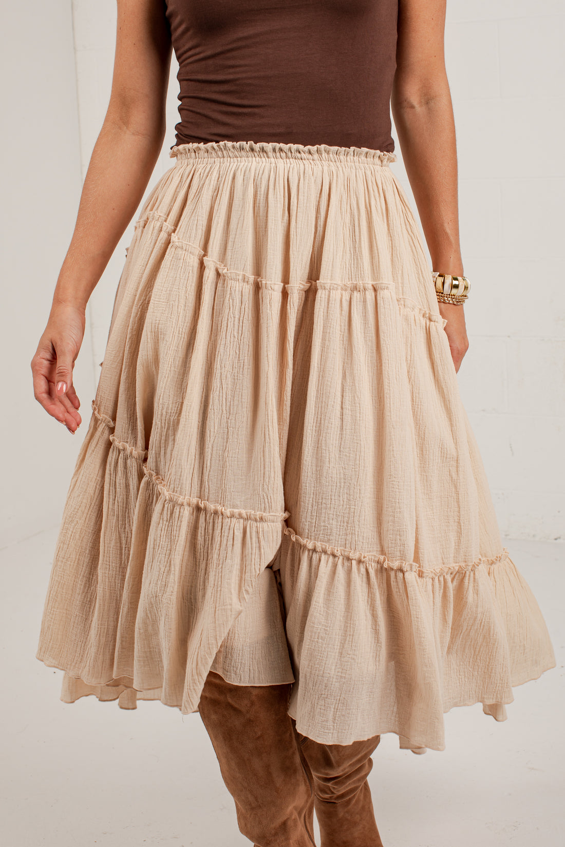 A beige tiered gauze skirt worn by a model, paired with a brown sleeveless top and brown boots, against a white wall.