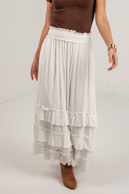 A model wearing a long white tiered maxi skirt with a smocked waistband, paired with a brown top and brown suede boots, shown from the chest down against a plain white studio background.