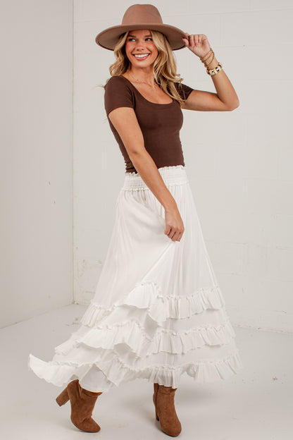 A woman modeling a bohemian-style outfit featuring a brown short-sleeve scoop-neck top, a long white tiered ruffle maxi skirt, a brown wide-brim hat, and brown suede ankle boots, set against a plain white studio background.