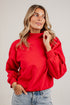 A woman wearing a red mockneck sweatshirt and light blue jeans, standing against a solid light gray background.