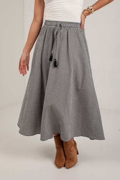 A woman wearing a black and white micro-checkered midi skirt with a drawstring waist, styled with a white top and brown suede ankle boots.