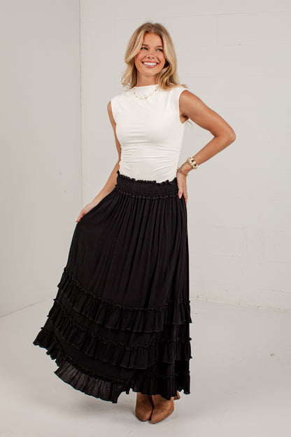 A woman modeling a long black tiered maxi skirt paired with a white sleeveless top, standing against a white studio background.