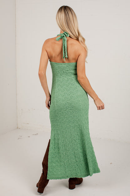 A woman shown from the back wearing a textured sage green halter-neck maxi dress with a tie at the back of the neck, paired with brown boots, in a white studio setting.