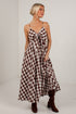 A woman modeling a brown and beige gingham maxi dress with spaghetti straps, a sweetheart neckline, and a front tie detail, paired with brown suede boots, set against a white brick wall background.