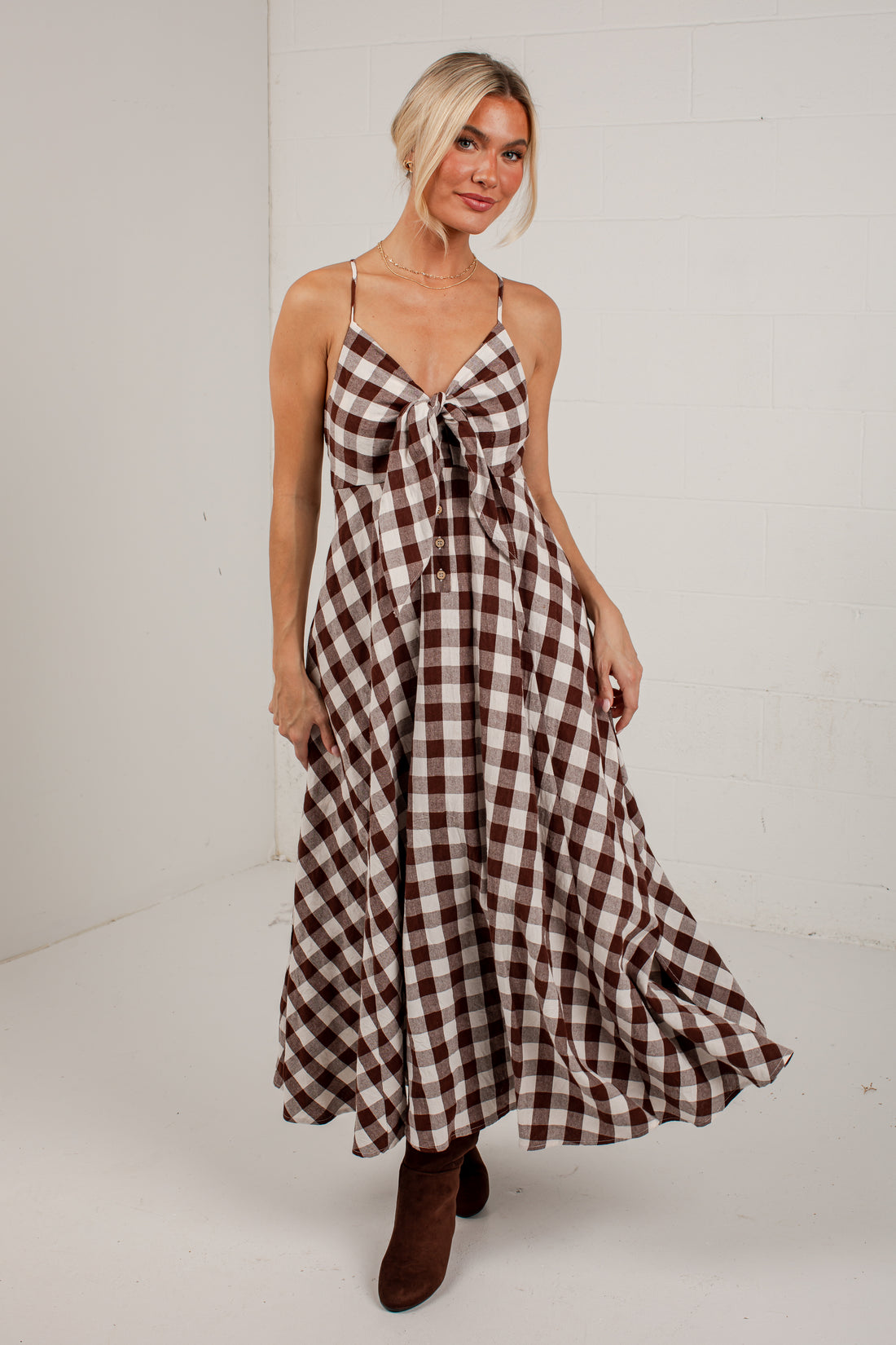 A woman modeling a brown and beige gingham maxi dress with spaghetti straps and a tie-front detail, paired with brown ankle boots, set against a white studio background.