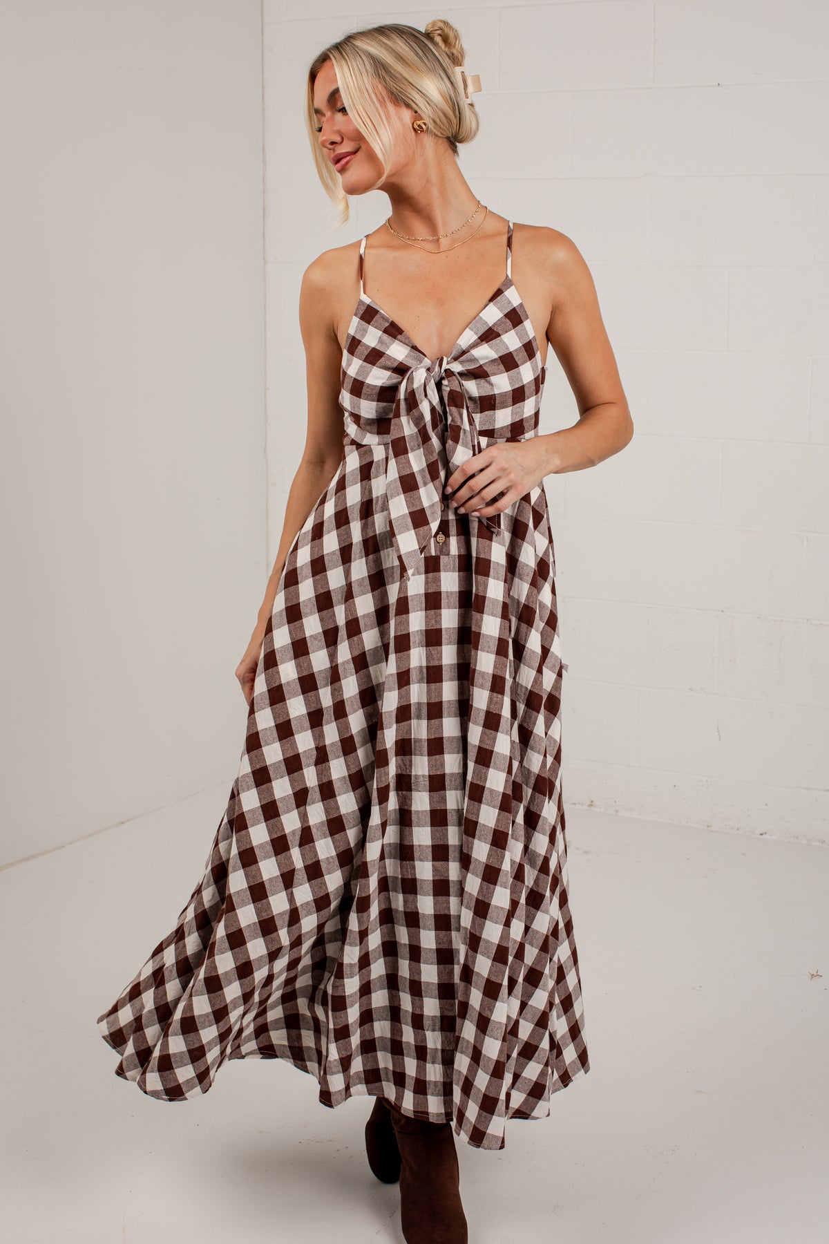 A woman modeling a brown and white gingham maxi dress with spaghetti straps and a tie-front detail, standing against a white studio wall.