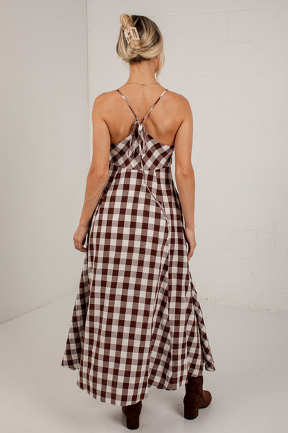 A woman viewed from behind wearing a brown and white gingham maxi dress with thin straps and a tie at the back, paired with brown boots, against a white studio background.