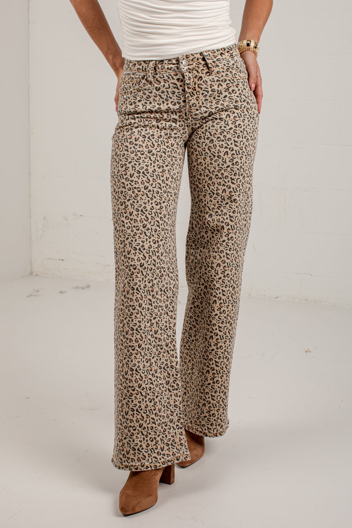 A person wearing leopard-print wide-leg pants with a white top and brown heeled shoes, standing against a plain light gray wall.