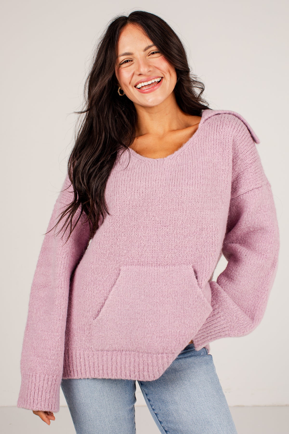 A woman smiling while wearing an oversized dusty pink knit sweater with shoulder details and a front kangaroo pocket, paired with light blue jeans, against a plain light gray background.