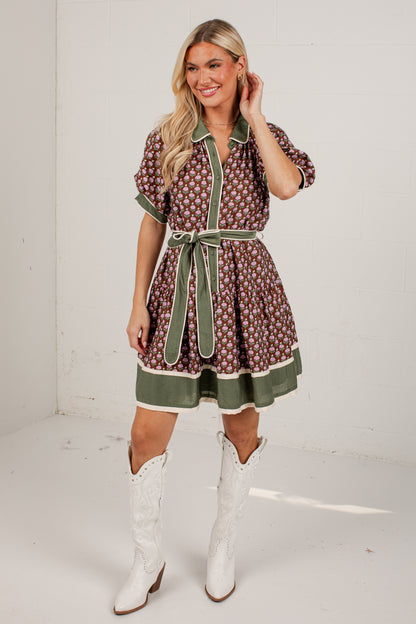 A woman modeling a short-sleeve brown and white floral print dress with sage green trim and a matching tie belt, paired with white cowboy boots, in a bright studio setting.