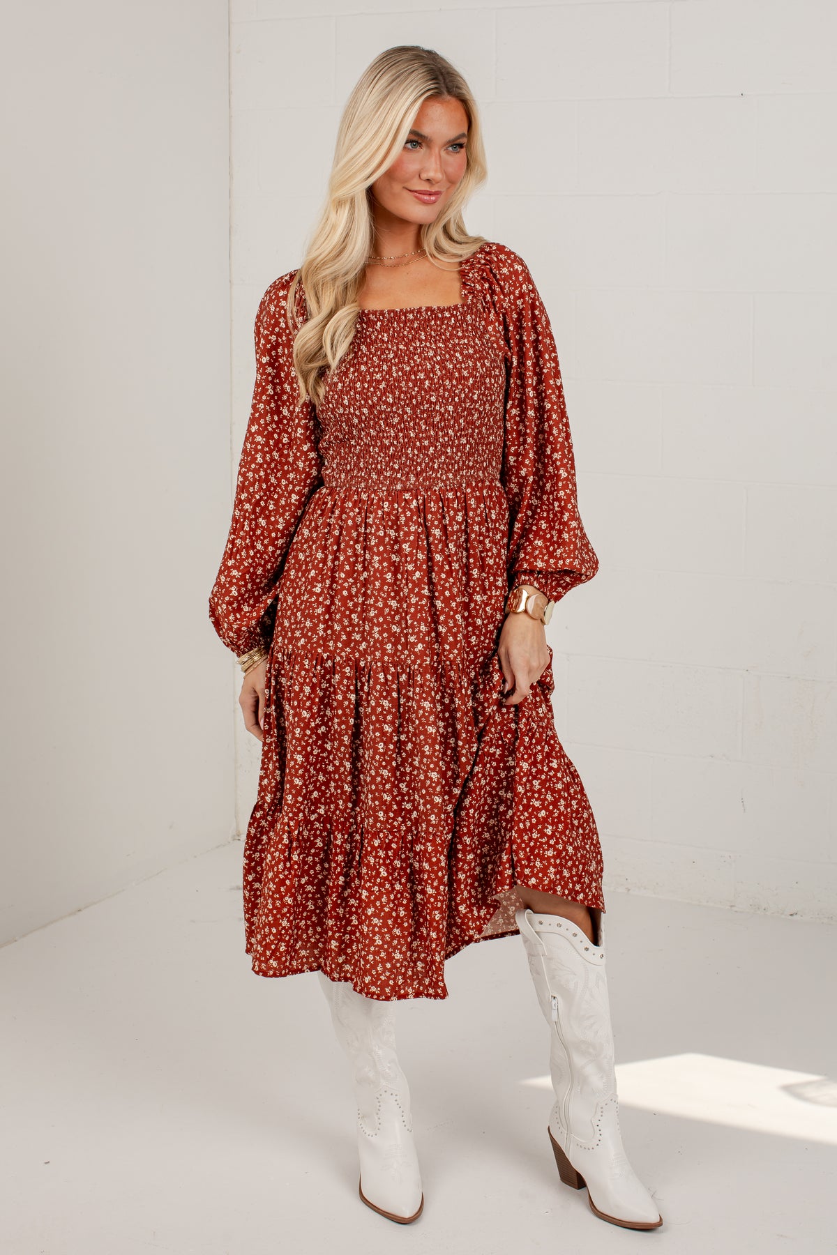A woman modeling a rust-colored, long-sleeved midi dress with a white floral print, paired with white cowboy boots, standing against a white studio background.