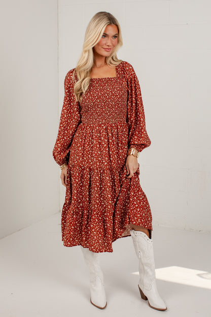 A woman modeling a rust-colored, long-sleeved midi dress with a white floral print, paired with white cowboy boots, standing against a white studio background.