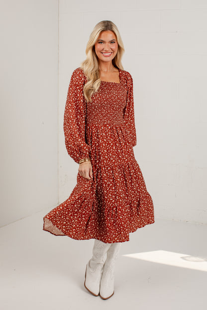 A woman modeling a rust-red long-sleeve midi dress with a white and orange floral pattern, paired with white knee-high boots, in a bright white studio setting.