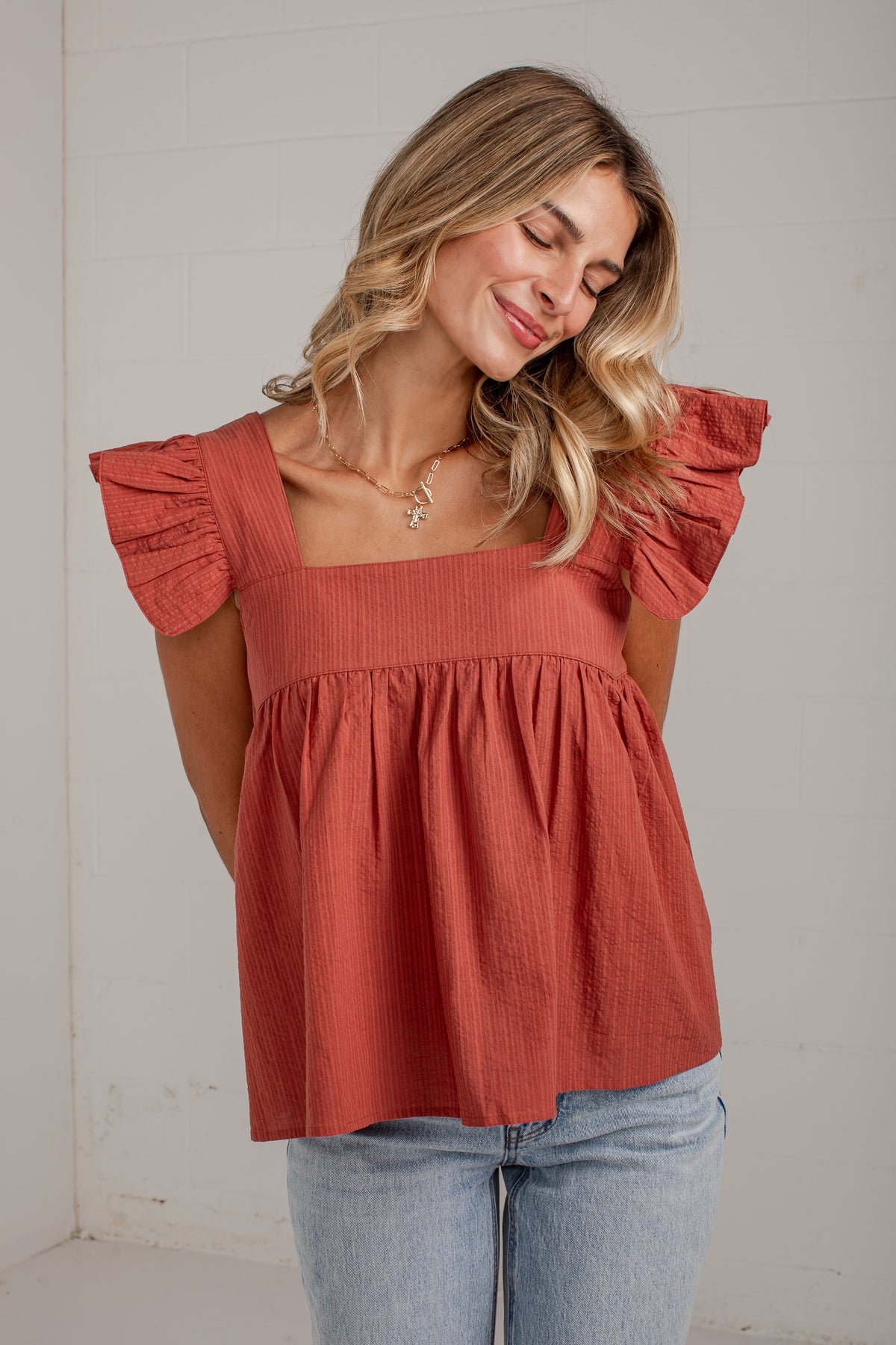 A woman wearing a terracotta-colored crinkle-textured top with ruffled sleeves and a square neckline, paired with light-wash blue jeans, standing against a white paneled wall.