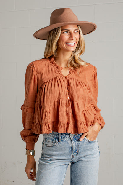 A woman wearing a terracotta orange tiered blouse with ruffled details, a brown wide-brim fedora hat, and light-wash blue jeans, standing against a white brick wall.