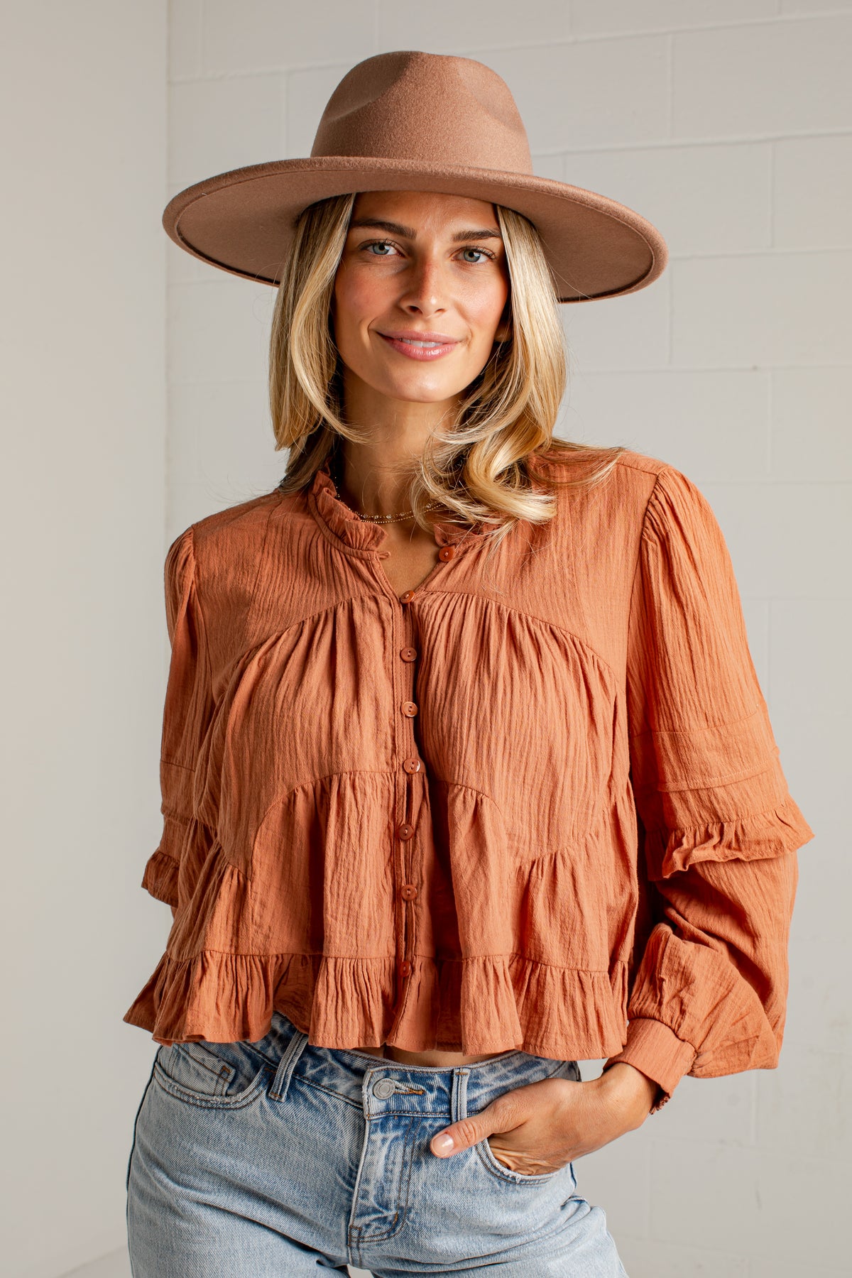 A woman wearing a terracotta-colored crinkled blouse with a ruffled collar and button front, paired with a wide-brimmed brown hat and light-wash blue jeans, standing against a white brick wall.