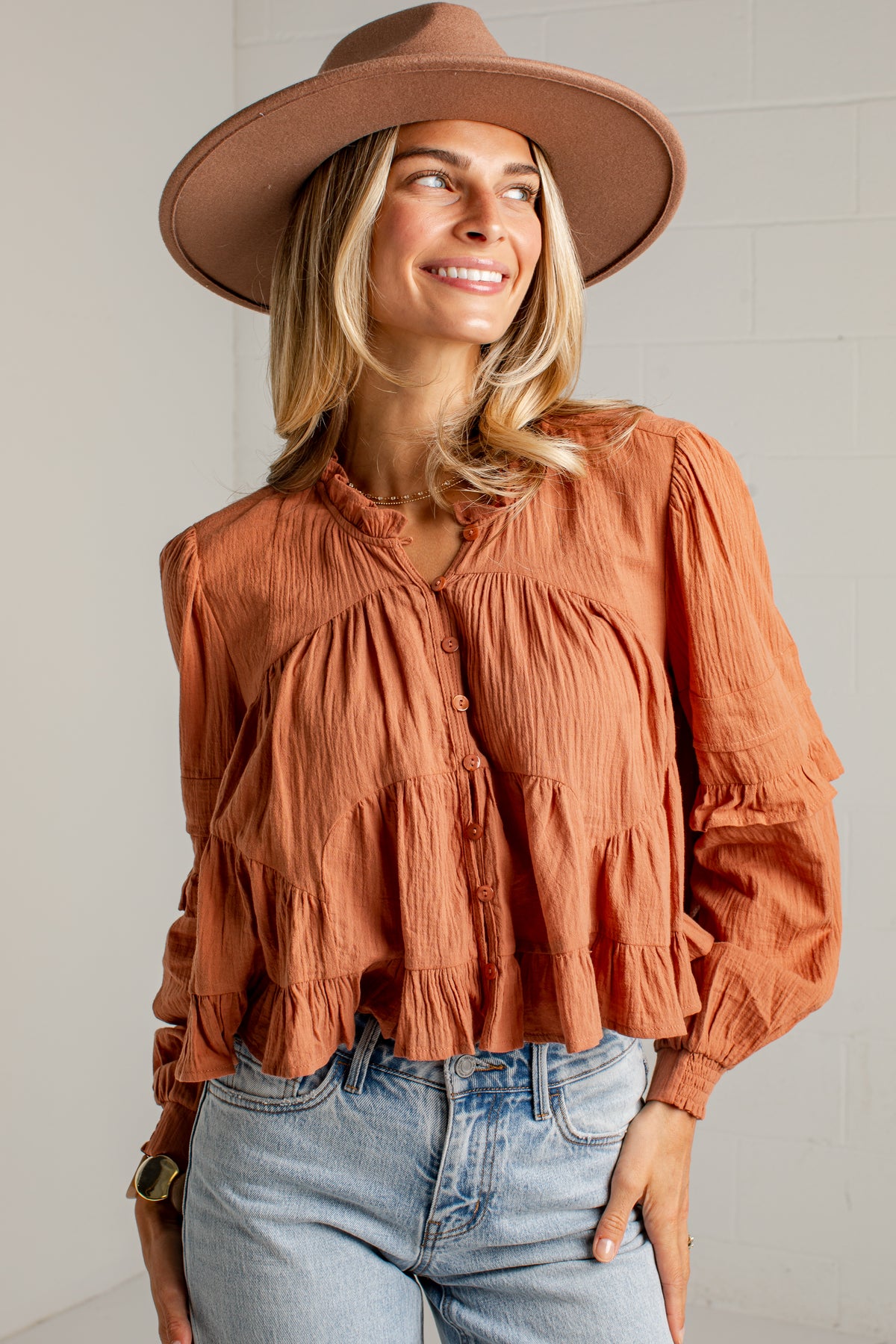 A woman wearing a terracotta orange crinkled long-sleeve blouse with a tiered ruffle hem, paired with light-wash blue jeans and a brown wide-brim hat, posing against a white brick wall.