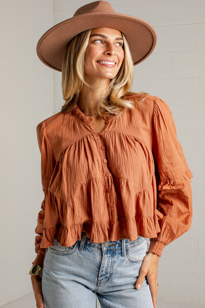 A woman wearing a terracotta orange crinkled long-sleeve blouse with a tiered ruffle hem, paired with light-wash blue jeans and a brown wide-brim hat, posing against a white brick wall.