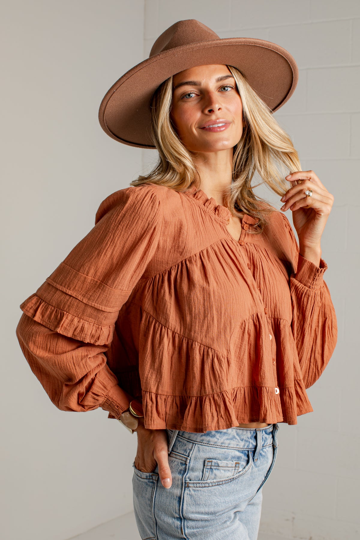 A woman wearing a terracotta-colored crinkle gauze blouse with tiered ruffles and long sleeves, paired with a brown wide-brim hat and light-wash blue jeans, standing against a white brick wall.