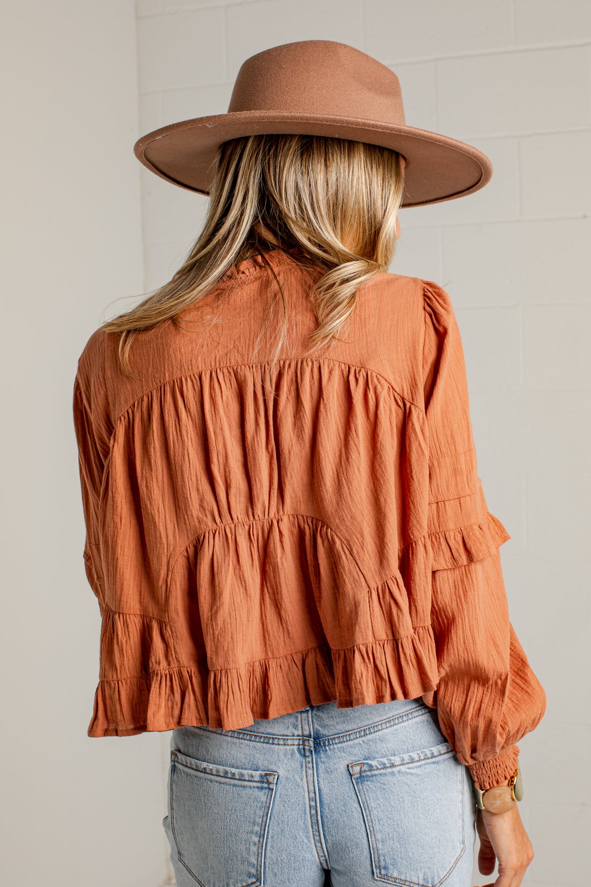 A woman viewed from behind wearing a terracotta orange ruffled blouse, a brown wide-brim hat, and light-wash blue jeans against a white brick wall.